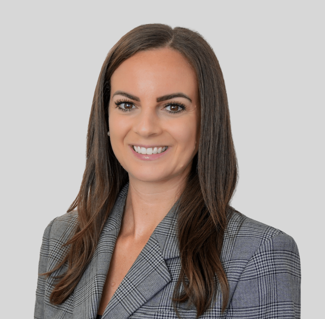 Professional headshot of a smiling woman with long brown hair, wearing a gray plaid blazer, against a light gray background.