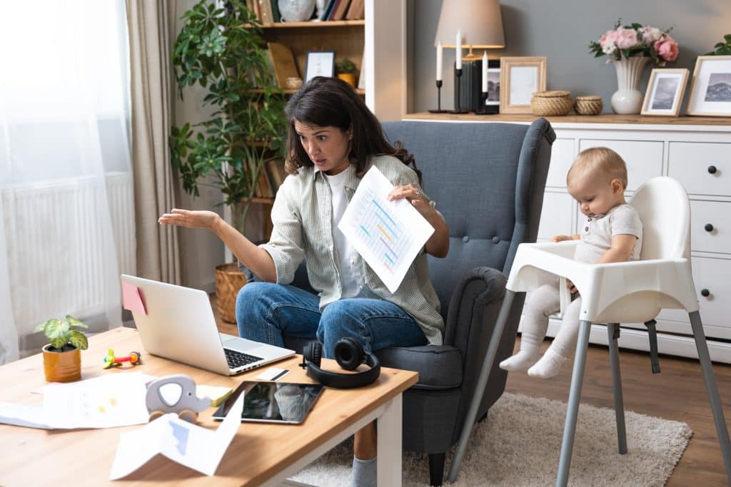 Woman sitting on a sofa, holding a printed chart while a laptop is open beside her, with a baby in a high chair nearby and a cluttered coffee table in a living room.